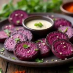 A beautifully plated purple sweet potato recipe featuring roasted wedges garnished with fresh herbs, accompanied by a creamy dipping sauce and lime wedges on a rustic wooden table.